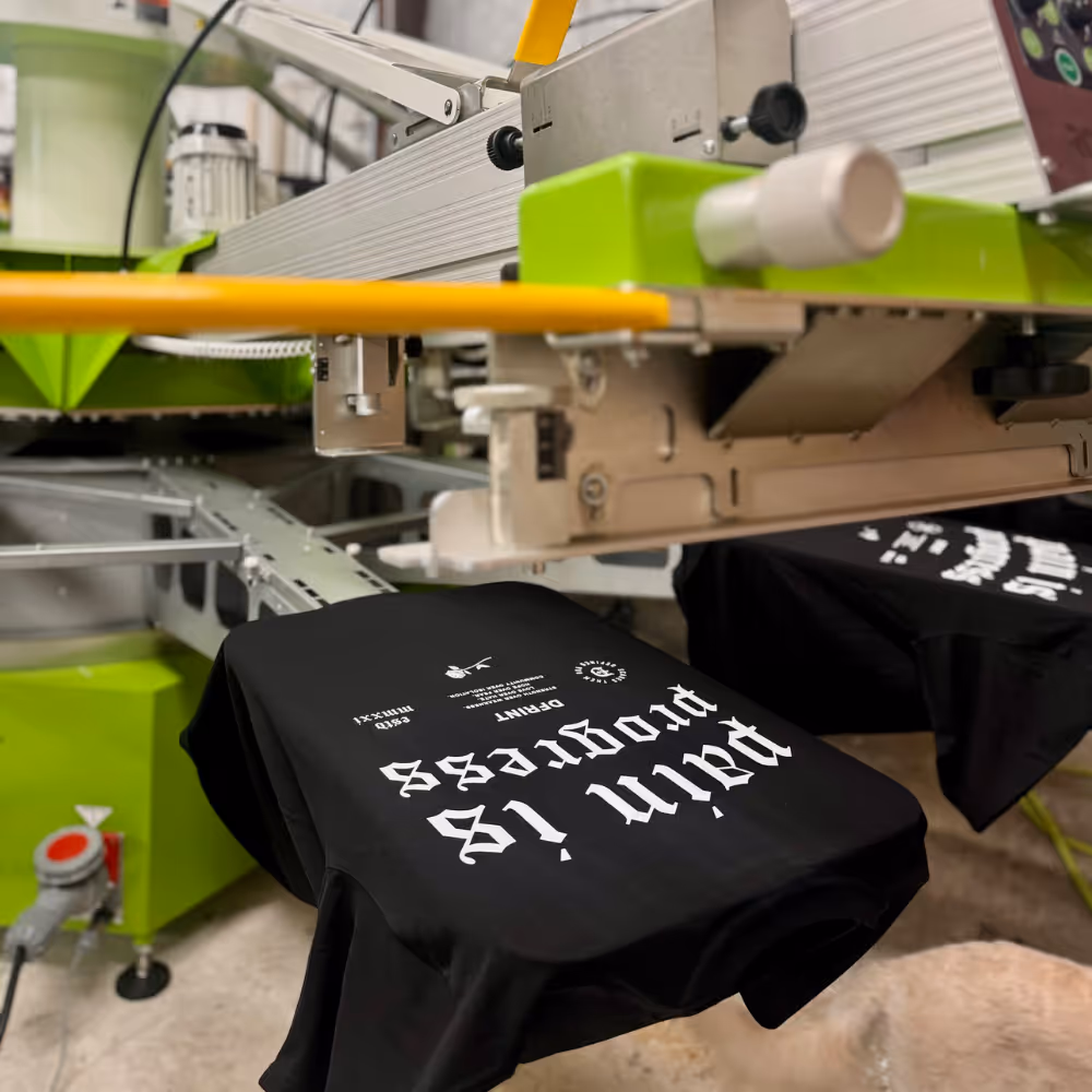 Black DFRINT t-shirts with 'Pain is Progress' slogan being screen printed on automatic press at Prophecy Print & Design.