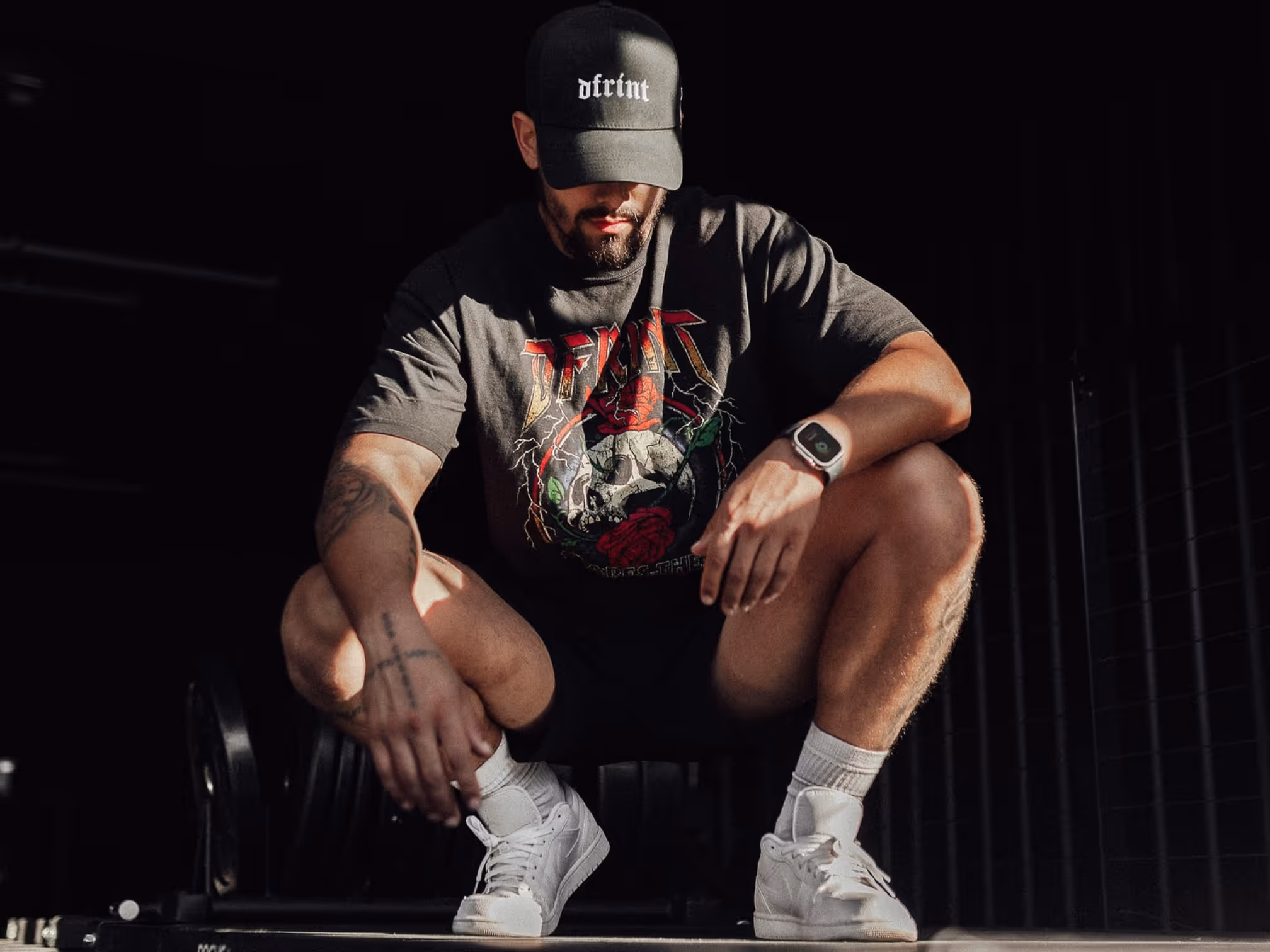 Man wearing DFRINT graphic t-shirt and hat, crouching in dim gym setting, showcasing streetwear apparel.