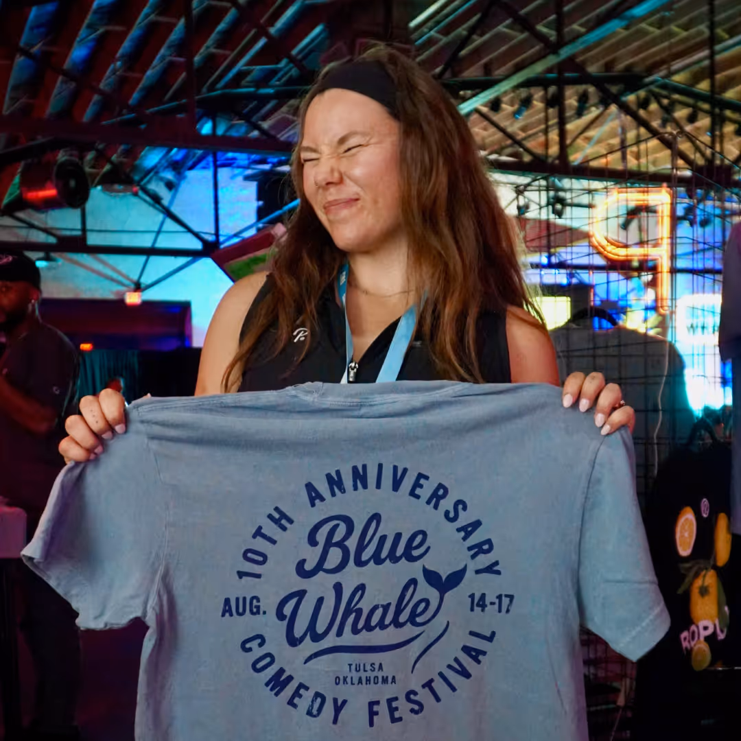 a girl holding up the 10th anniversary blue whale comedy festival live printed shirt