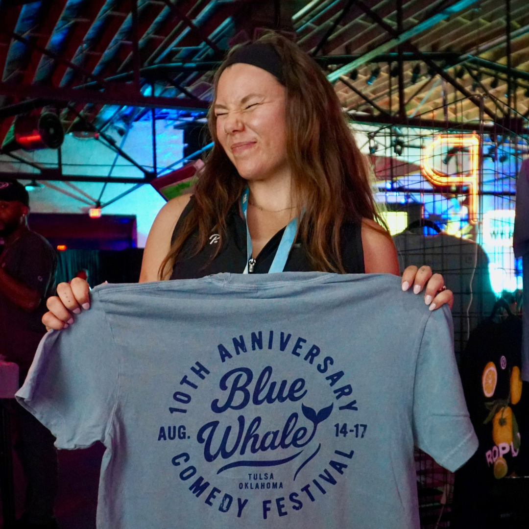 a girl holding up the 10th anniversary blue whale comedy festival live printed shirt