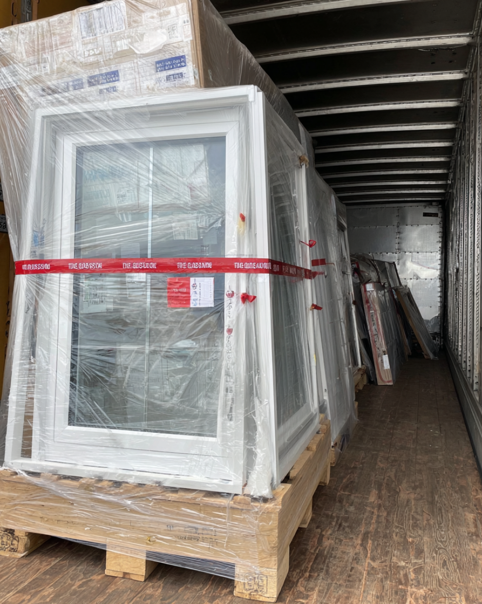 Large glass windows on pallets wrapped in plastic film, secured with red tape inside a truck for transport.