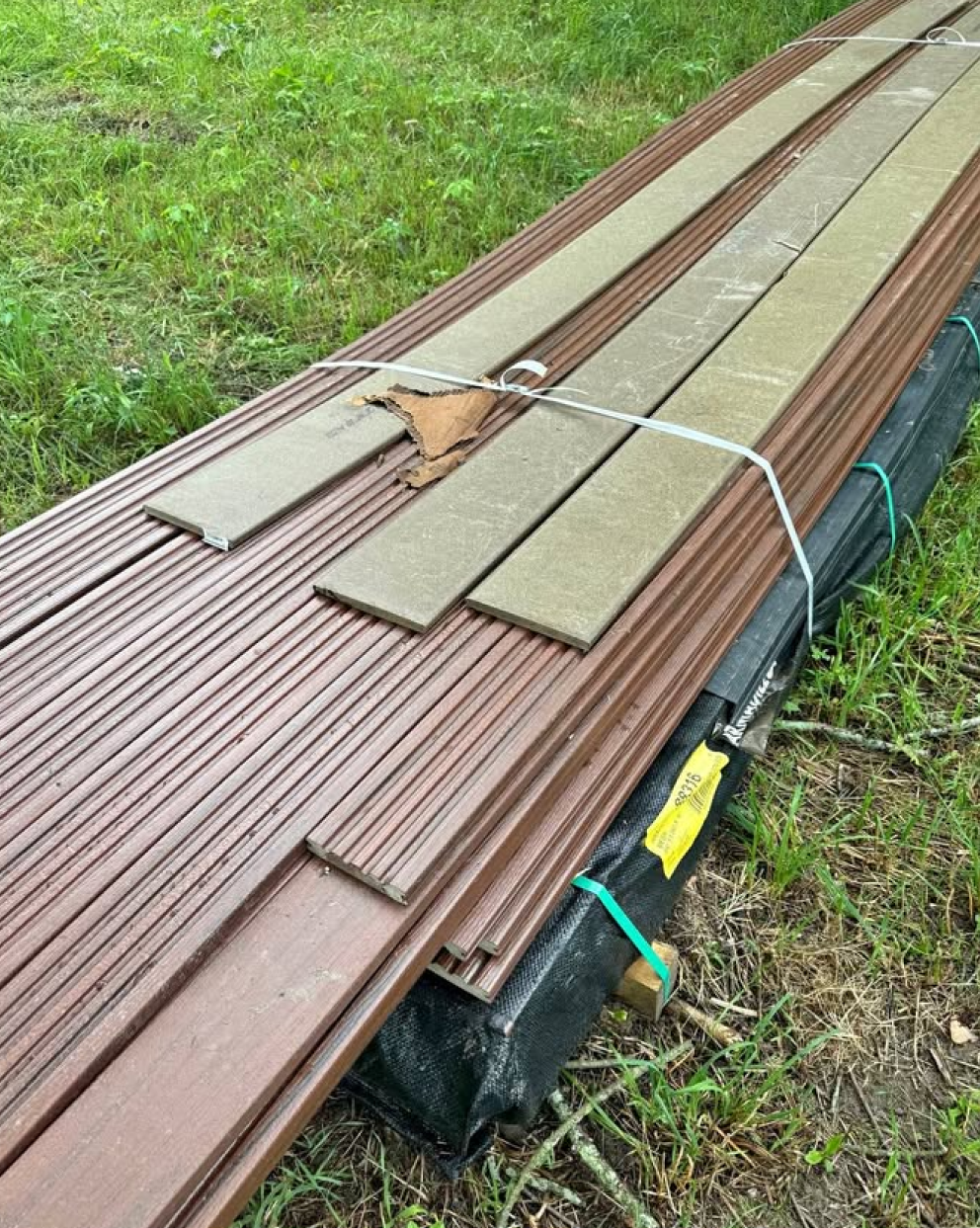 Weathered brown composite decking boards with straps – Bundle of older brown composite deck boards tied together with straps, placed on grass.