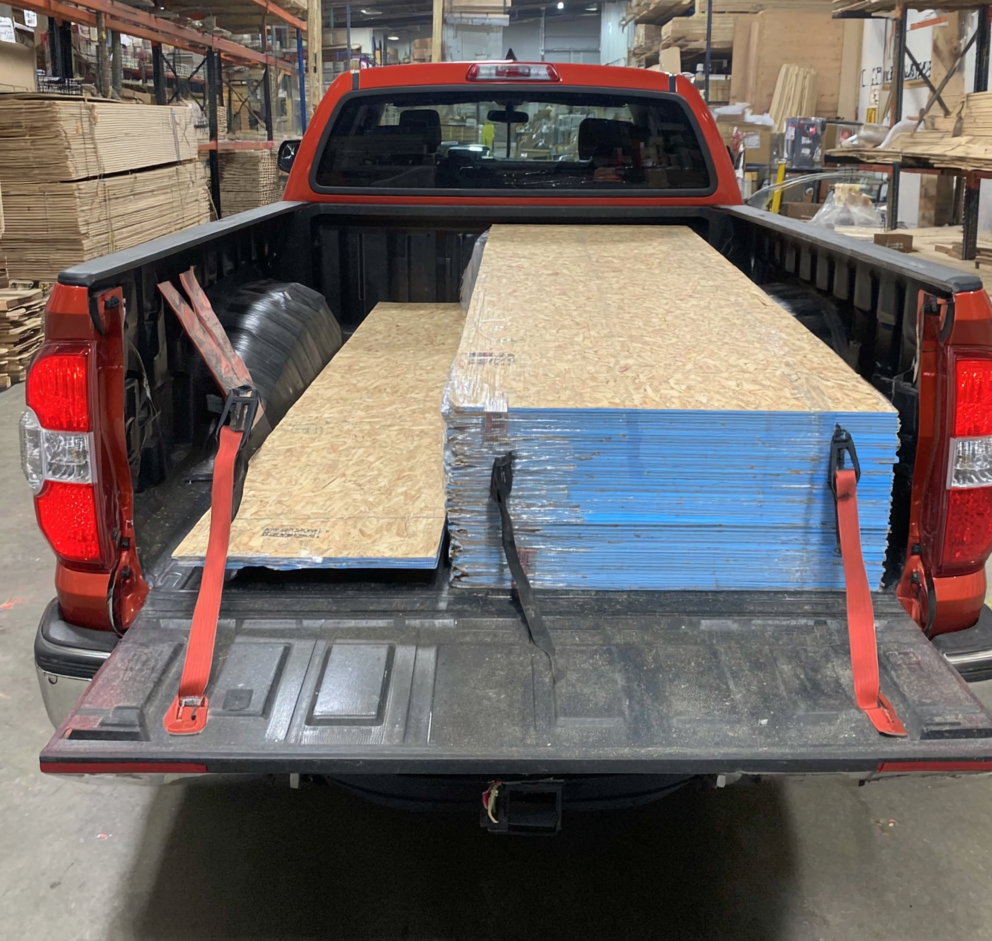 Stack of plywood sheets loaded in a red pickup truck bed inside a lumber warehouse, secured with straps