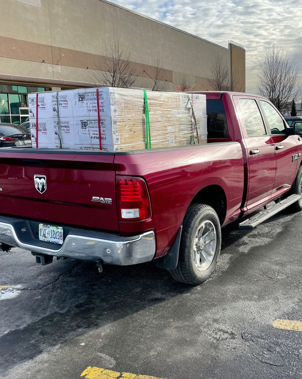 A red Dodge pickup truck carrying a pallet of shrink-wrapped flooring boxes secured with green straps in its bed, parked in a lot outside a large store.