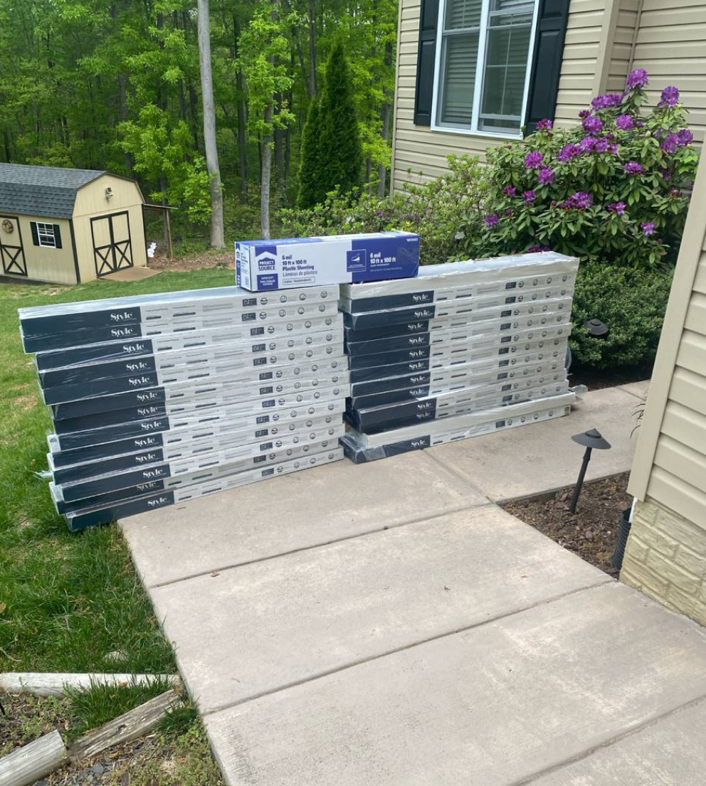 Multiple stacks of black and white packaged flooring branded “Style Selections” neatly arranged outside a house, next to a garden with flowers and greenery.