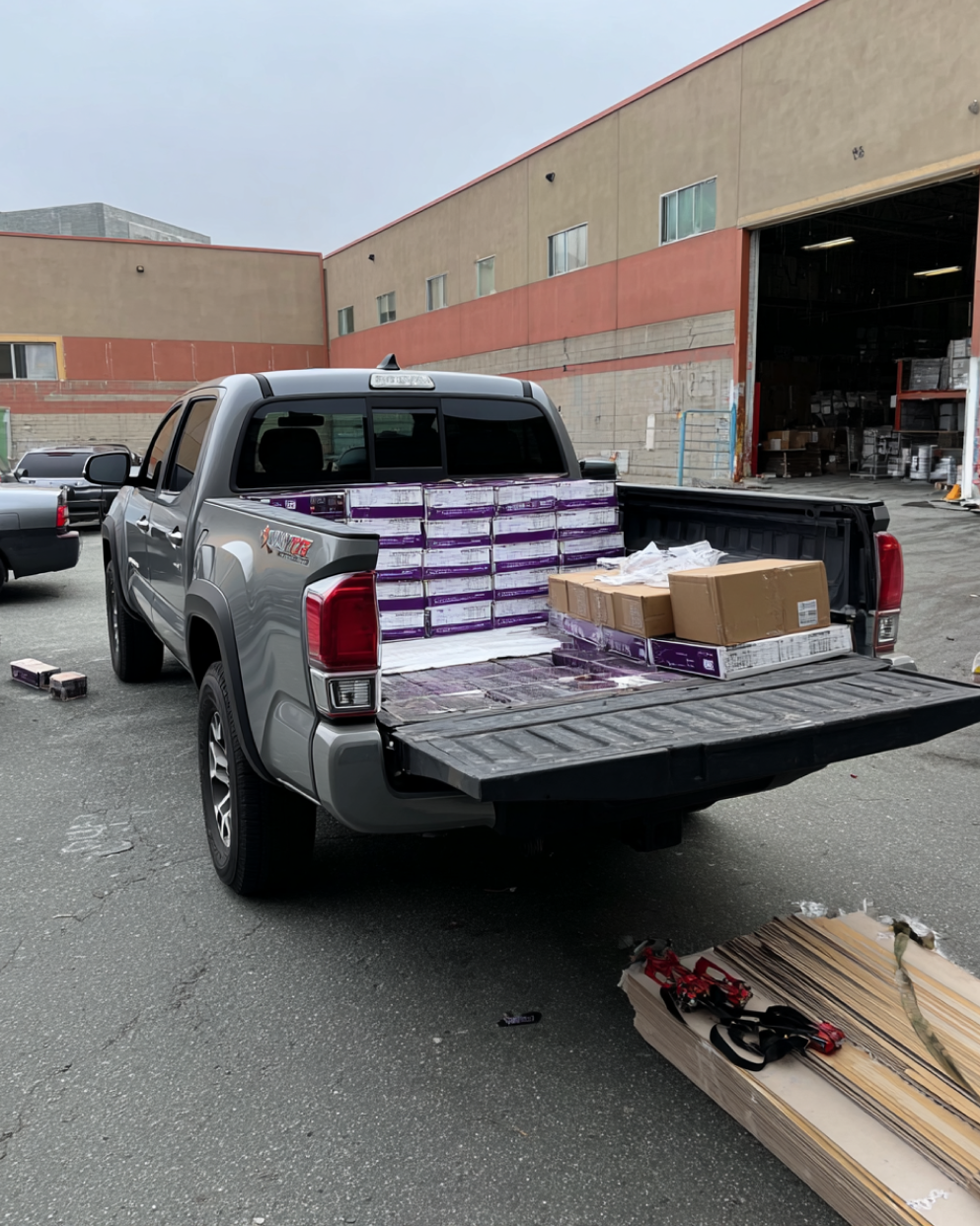 A gray pickup truck bed loaded with purple-boxed flooring packs and a few brown boxes, parked outside a warehouse with scattered flooring pieces on the ground nearby.