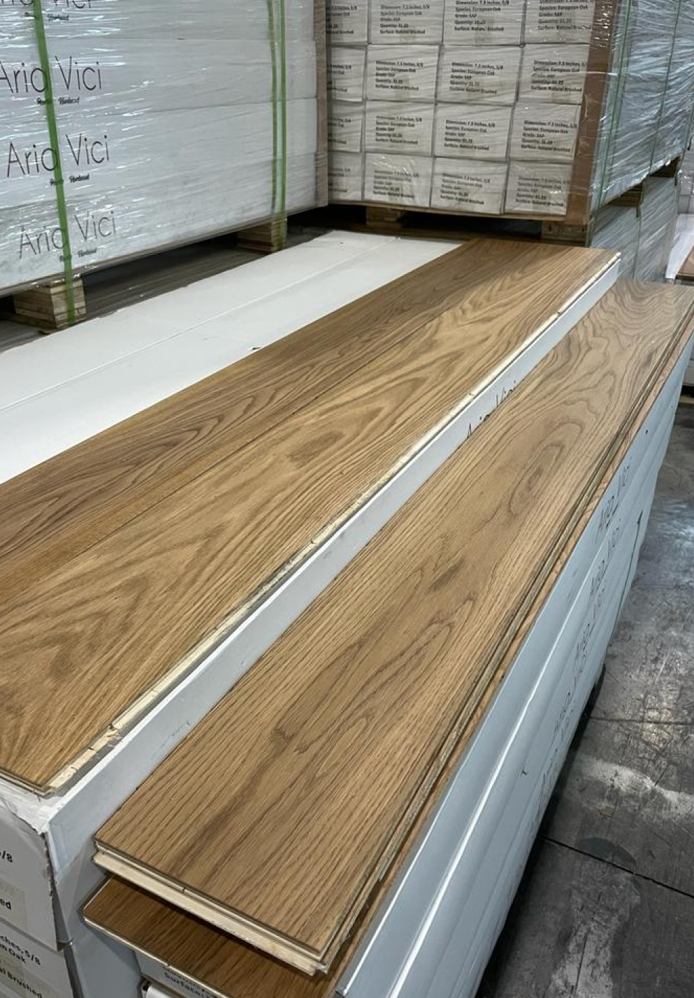 A stack of light brown wooden flooring planks displayed on top of white packaging in a warehouse, with other pallets of flooring visible in the background.