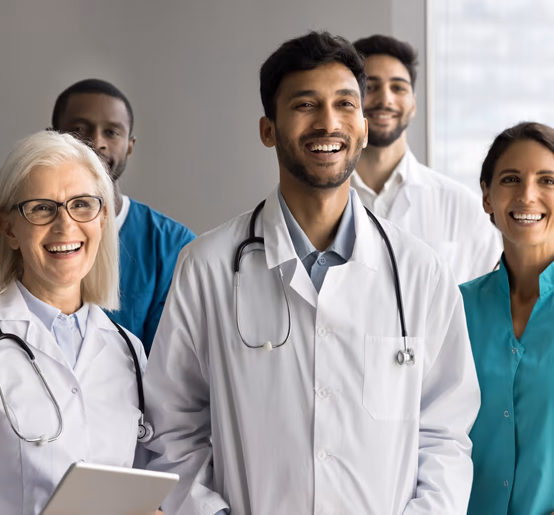 A diverse group of smiling healthcare professionals standing together, including doctors in white coats with stethoscopes and a nurse in teal scrubs, conveying teamwork and positivity in a medical setting.