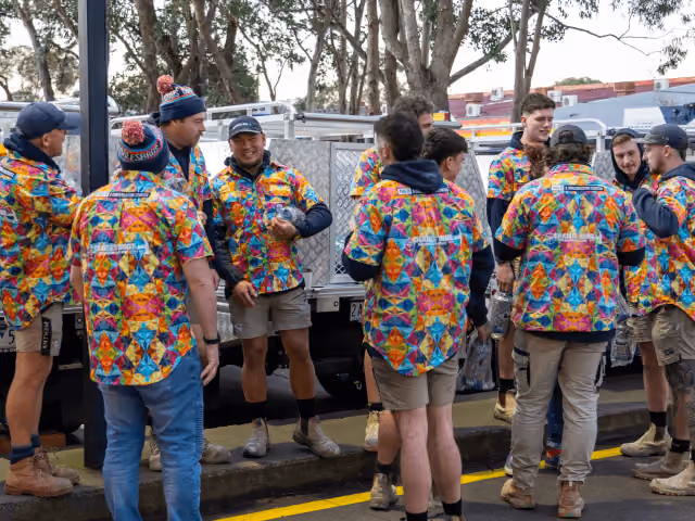 Group of people wearing colorful patterned shirts talking near vehicles on a street with trees.