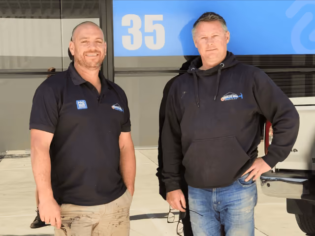 Two men in casual work attire standing outside a building with number 35 on a blue sign.