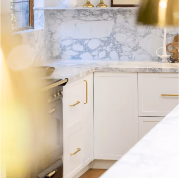 Bright kitchen corner with white marble countertops, brass handles, and a black stove next to a window.