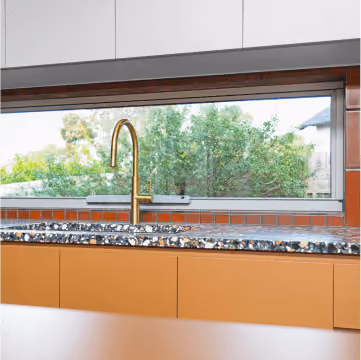 Modern kitchen sink with gold faucet below a long window overlooking green trees and a fence.