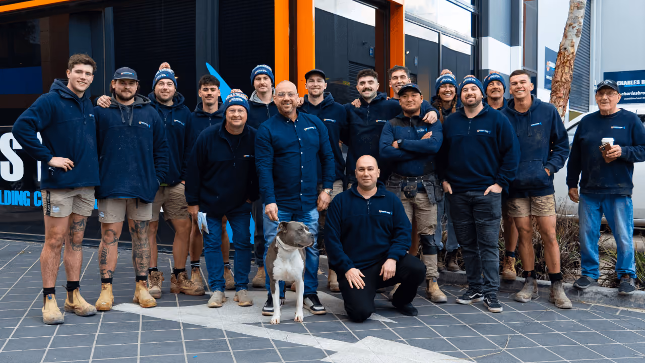 A group of workers in navy jackets posing outside a building with a dog in front.