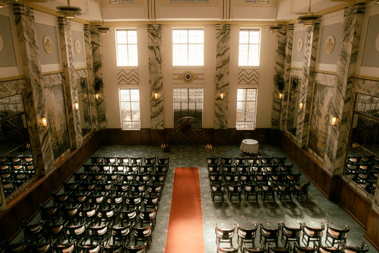 A grand indoor ceremony space with rows of dark wooden chairs arranged along a central red aisle, set in an elegant hall with marble columns, tall windows, and warm golden lighting.