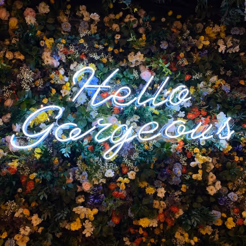 A neon sign reading “Hello Gorgeous” glowing in white light against a lush floral wall filled with colorful flowers and greenery.