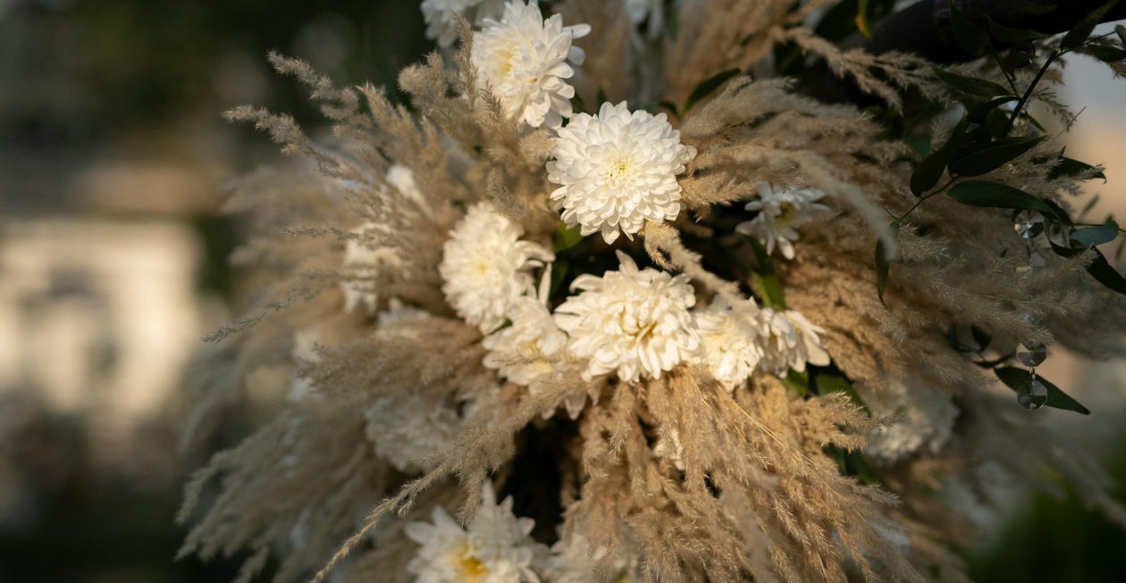 A close-up of a floral arrangement featuring soft beige pampas grass and white chrysanthemums against an outdoor background.