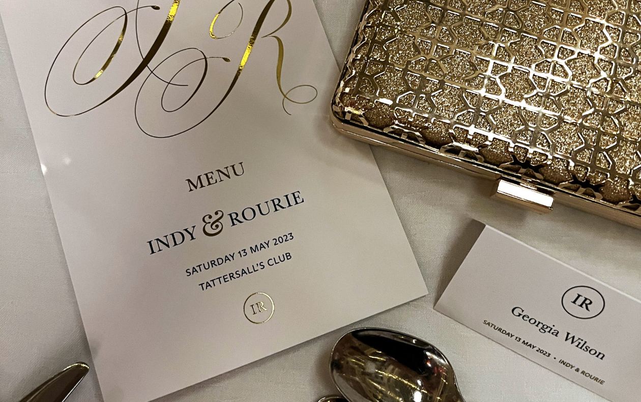 A close-up of an elegant wedding place-setting featuring a printed menu titled “Indy & Rourie,” dated 13 May 2023 at Tattersall’s Club, alongside a guest name card for Georgia Wilson, silver cutlery, and a gold textured clutch.