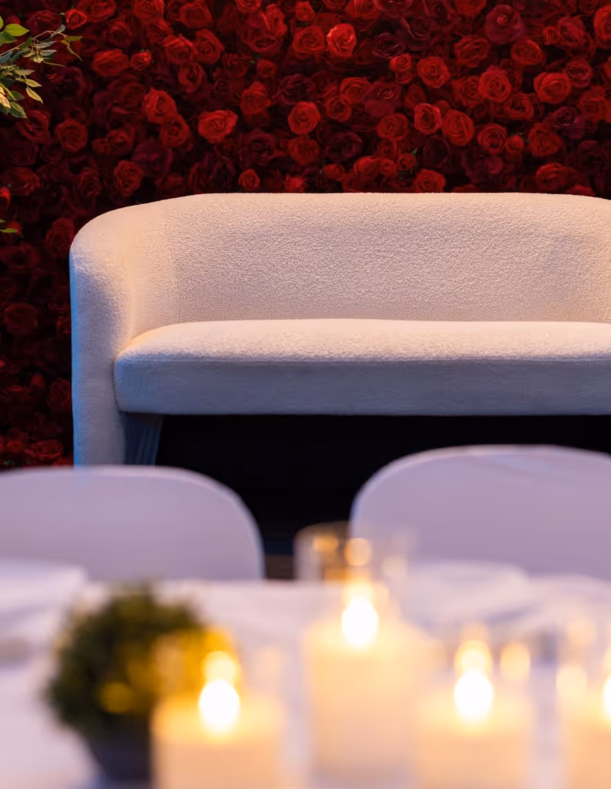 White curved sofa against a wall of red roses and blurred candles in the foreground.