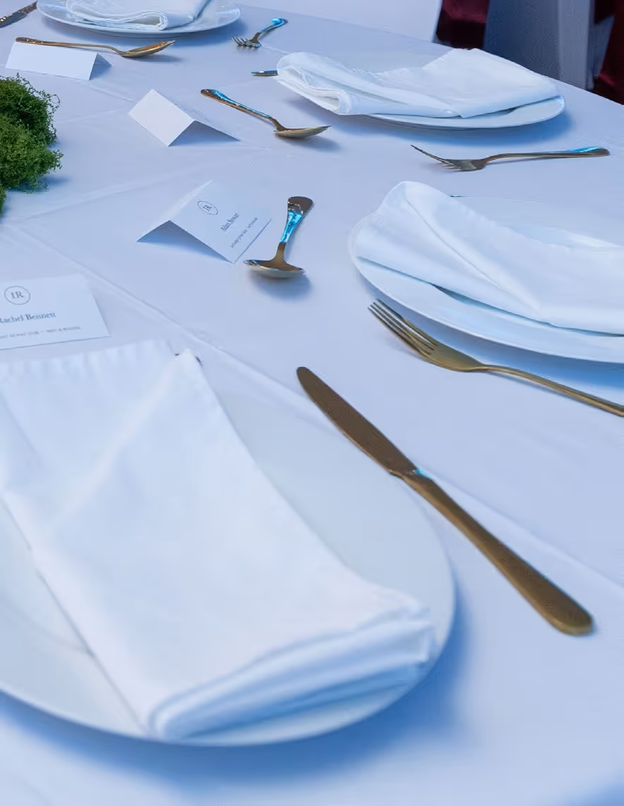 Formal table setting with white plates, folded napkins, gold cutlery, and name cards on a white tablecloth.