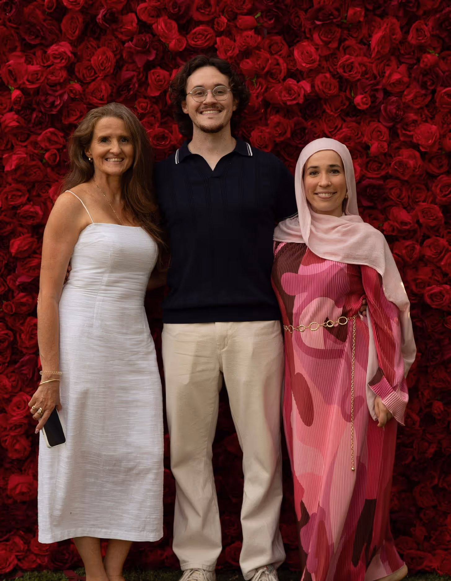 Three people smiling in front of red rose wall: woman in white dress, man in dark shirt, woman in pink hijab.