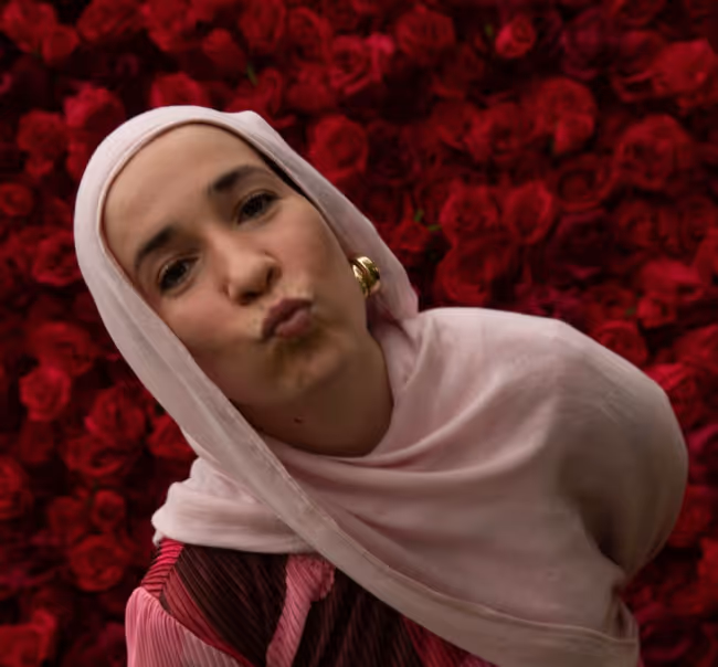 Woman in light pink hijab and gold earrings making a kissing face in front of red roses.