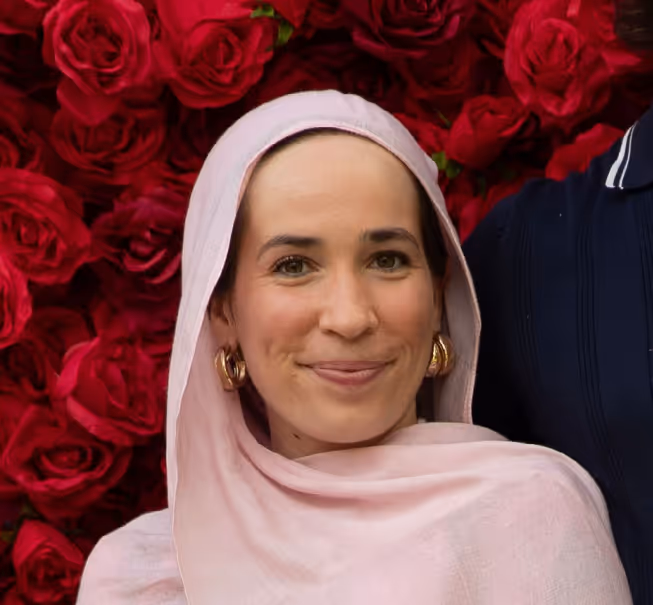 Smiling woman wearing a light pink headscarf and gold hoop earrings with red roses background.