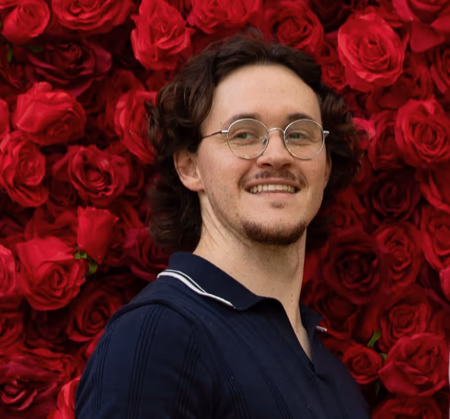 Smiling man with glasses and curly hair stands in front of a vibrant red rose wall.