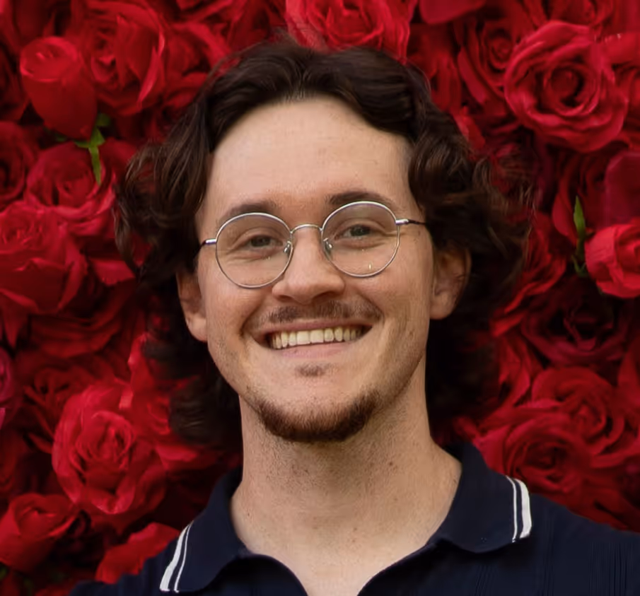 Smiling man with glasses and curly hair in front of a wall covered with red roses.