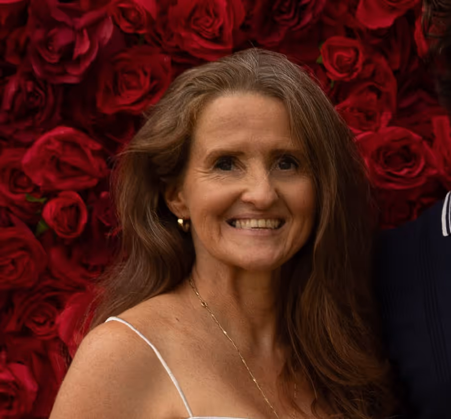 Smiling woman with long brown hair in white dress in front of a red rose flower wall.