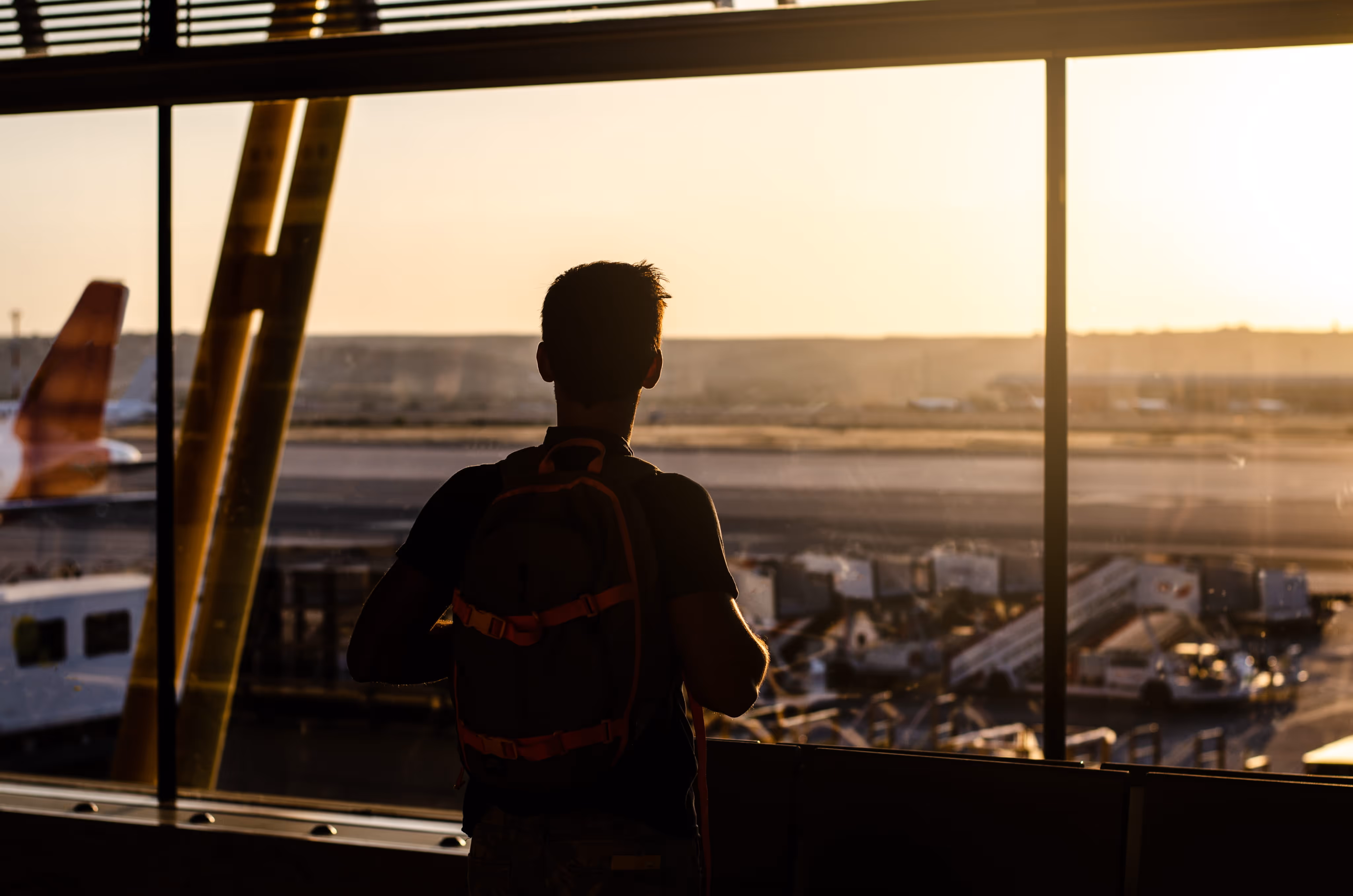 Silhouette of a person with a backpack looking out an airport window at a runway during sunset.