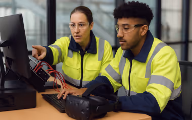 Two engineers in high-visibility jackets working with a computer and virtual reality headset in an office.