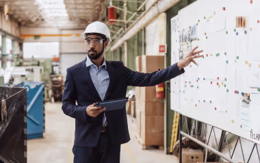 Man in a suit and safety helmet pointing at notes on a whiteboard inside an industrial workspace.