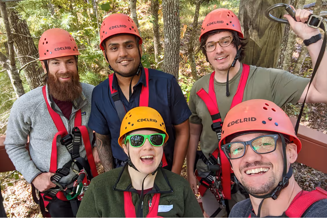 Group of five ArborXR employees wearing helmets and safety harnesses outdoors, smiling at the camera in a wooded area. Ready to go ziplining