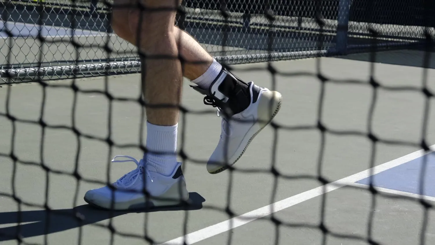 Close-up of a person wearing white tennis shoes and an ankle brace on a tennis court seen through a net.