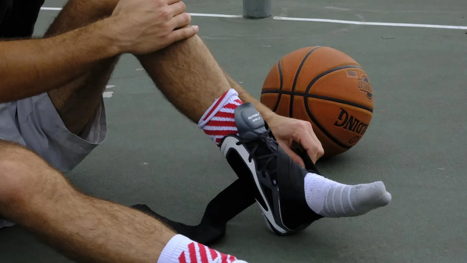 Person sitting on a basketball court putting on an ankle brace over a basketball shoe with a basketball nearby.