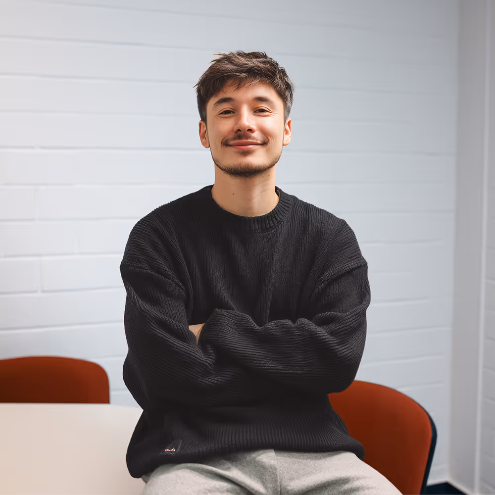 Ein junger Mann in einem schwarzen Pullover sitzt lächelnd an einem Tisch, im Hintergrund eine weiße Wand mit Kalender.