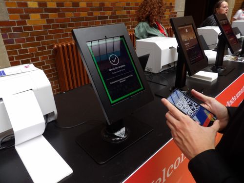 Een zelfbedienings check-in station met meerdere zwarte tablets en printers. Een deelnemer scant een QR-code vanaf een smartphone om toegang te krijgen. Het scherm van de tablet toont een bevestiging. Andere bezoekers gebruiken ook de apparaten, wat wijst op een efficiënte en digitale check-in oplossing.