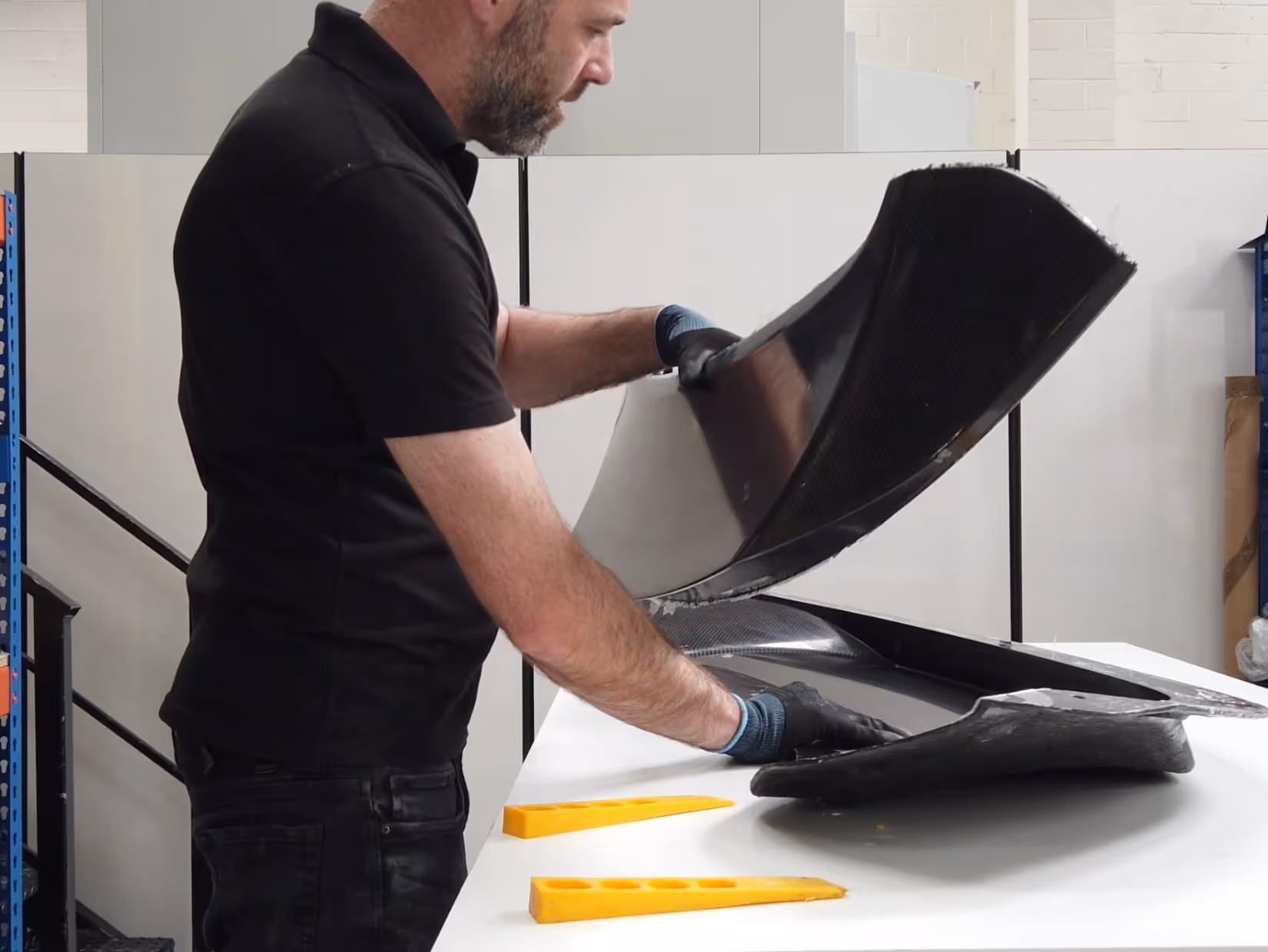 Man wearing black gloves examining large curved black composite material on a white table in a workshop.