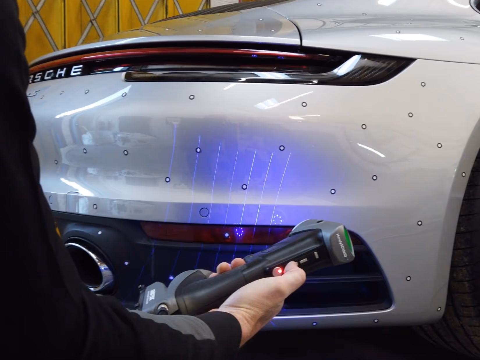 Person holding a handheld scanner with blue lights scanning the rear bumper of a silver Porsche car.