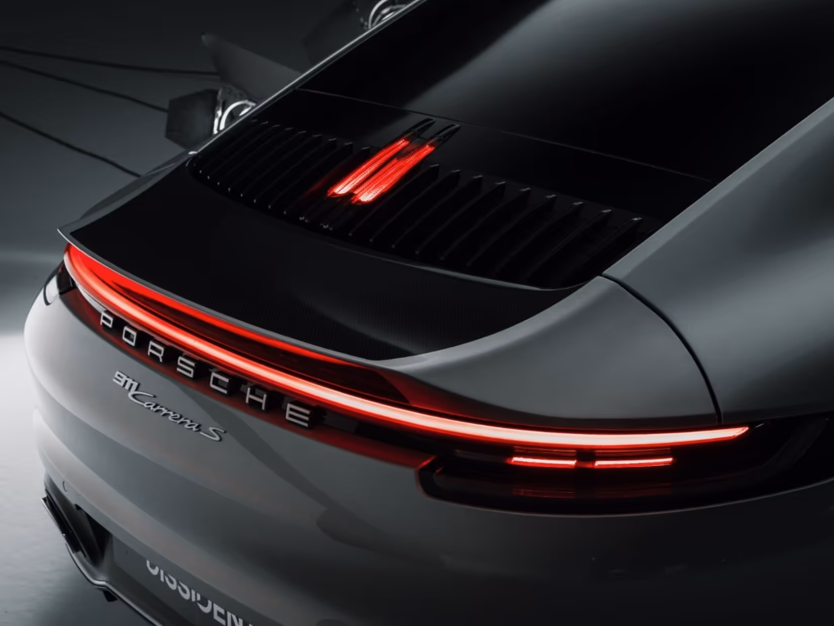 Rear view of a gray Porsche 911 Carrera S showing illuminated taillights and the Porsche emblem.