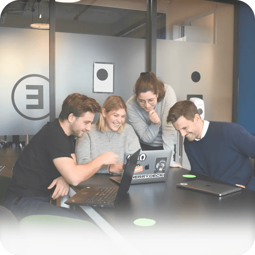 Four colleagues smiling and collaborating around two laptops in a modern office meeting room.