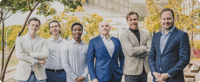 Six men in business casual and formal attire standing outdoors with trees and a building in the background.