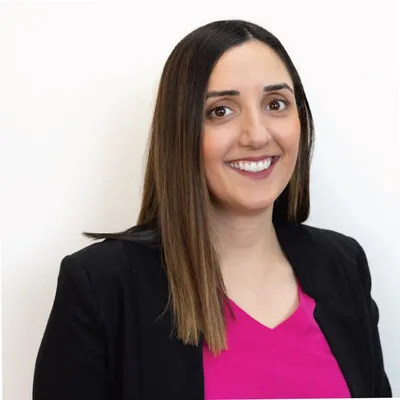 Smiling woman with long straight brown hair wearing a black blazer and pink top against a white background.