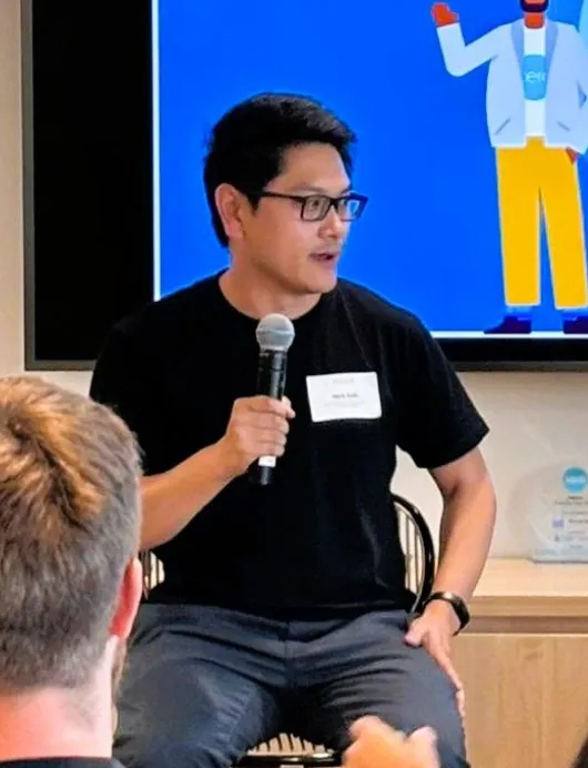 Man wearing glasses and a black shirt holding a microphone and speaking during a meeting or presentation.