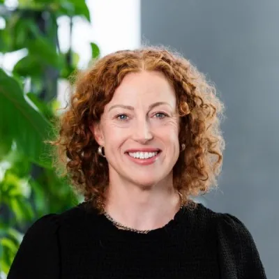 Smiling woman with curly red hair wearing a black top, standing in front of a blurred background with green plants.