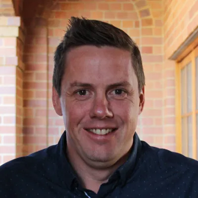 Smiling man with short dark hair wearing a dark blue shirt standing in front of a brick wall with arches.