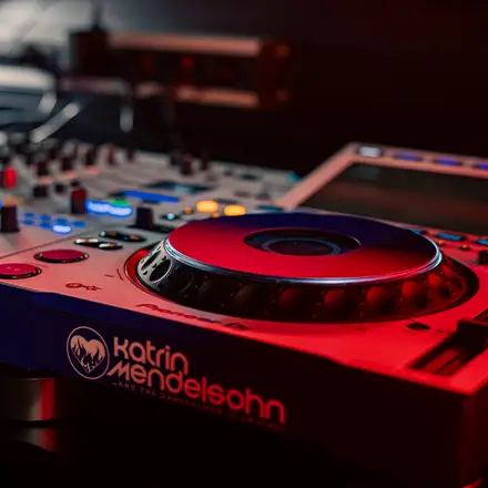 Detailaufnahme eines beleuchteten DJ-Pults mit Branding „Katrin Mendelsohn“ – in Rotlicht gehalten.