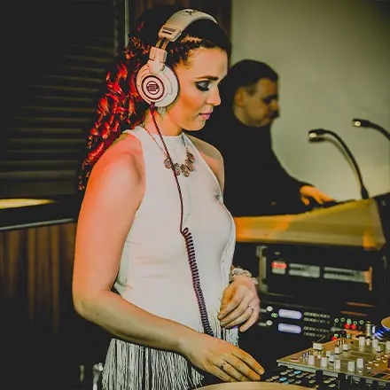DJane Katrin Mendelsohn steht am DJ-Pult bei einer stilvollen Hochzeit und legt konzentriert Musik auf, im Hintergrund ein Musiker am Flügel.