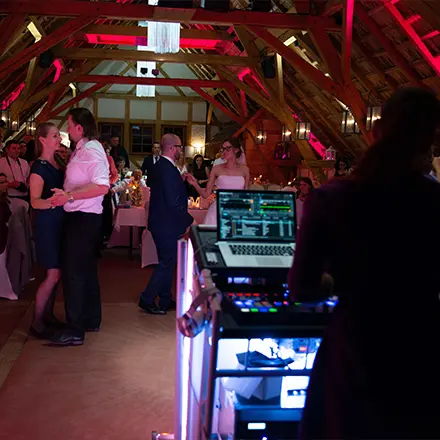 Blick vom DJ-Pult auf eine tanzende Hochzeitsgesellschaft in einer romantisch beleuchteten Eventlocation mit Holzdachkonstruktion.
