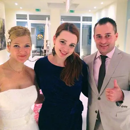 DJane Katrin Mendelsohn steht lächelnd zwischen einem frisch verheirateten Brautpaar. Die Braut trägt ein weißes Hochzeitskleid, der Bräutigam einen beigen Anzug mit Krawatte und zeigt einen Daumen hoch.